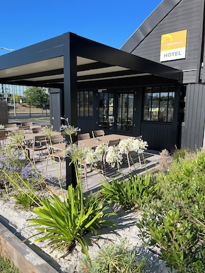 A hotel with an outdoor seating area, covered by a roof, and surrounded by plants. - The Originals Access, Hôtel Bordeaux Aéroport (Merignac)