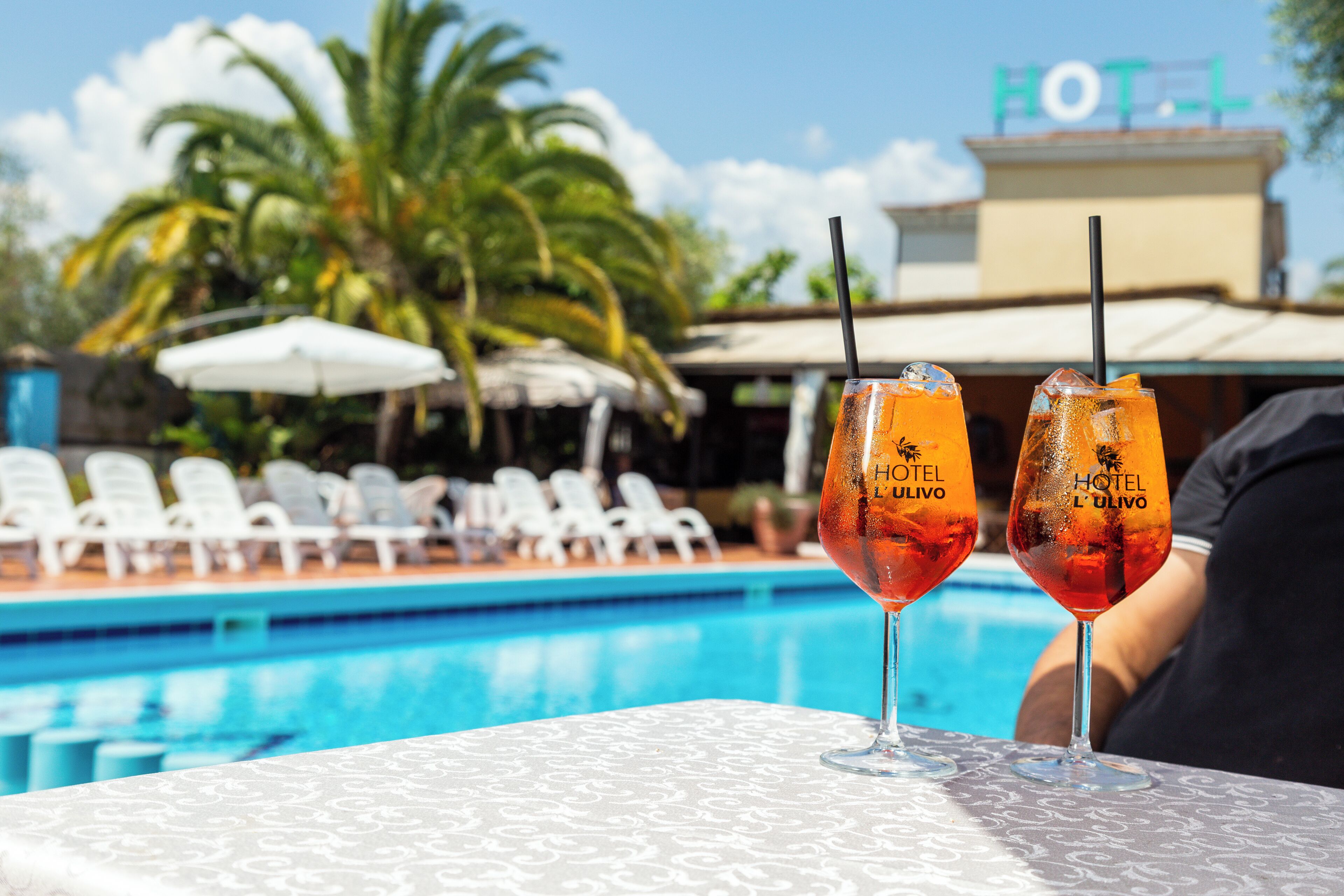 Deux cocktails sur une table près d’une piscine, avec des chaises longues et une enseigne d’hôtel en arrière-plan.