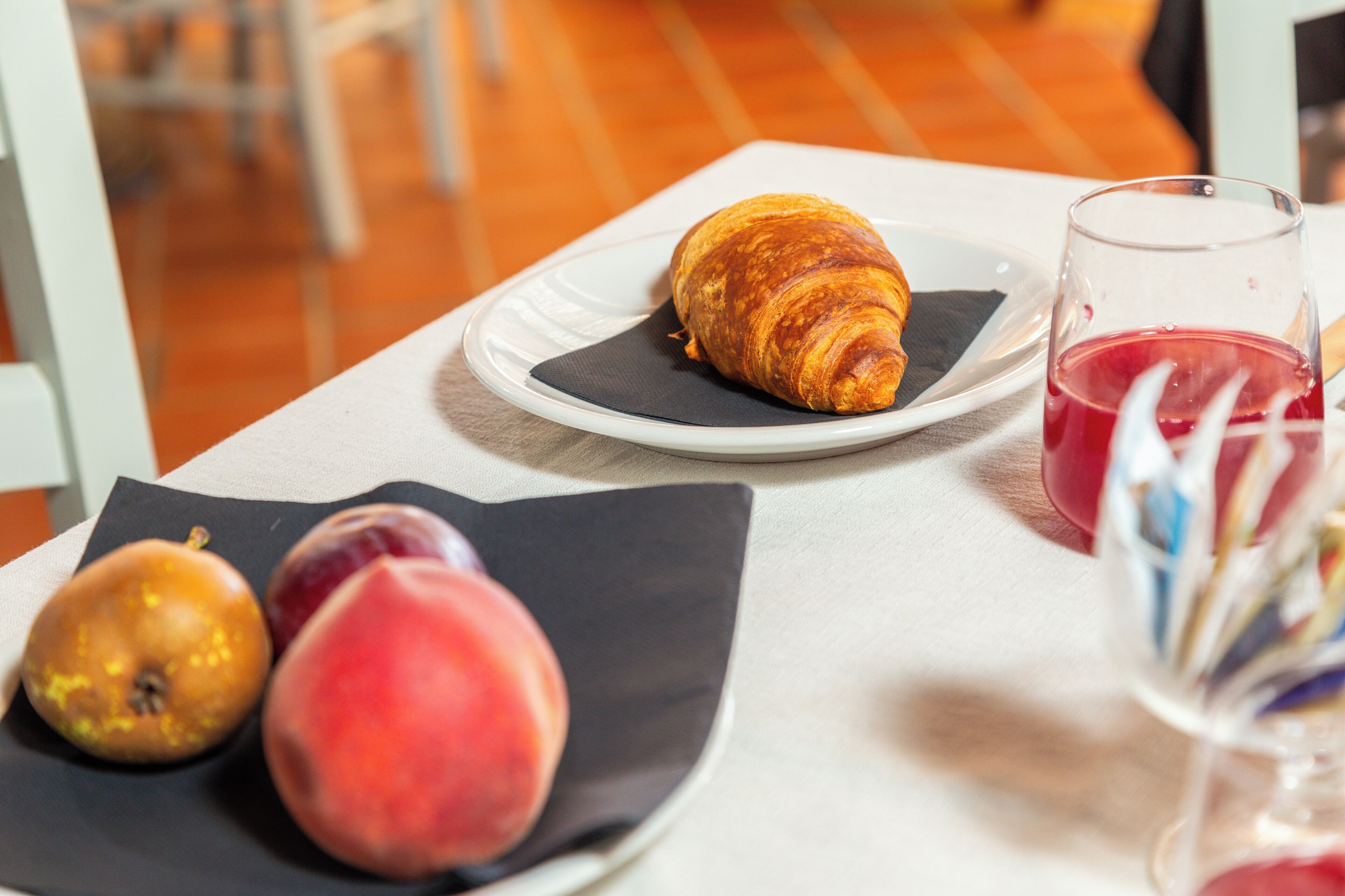 Ein Tisch mit einem Croissant, einem Glas roter Flüssigkeit und zwei Früchten auf einem Servietttuch.