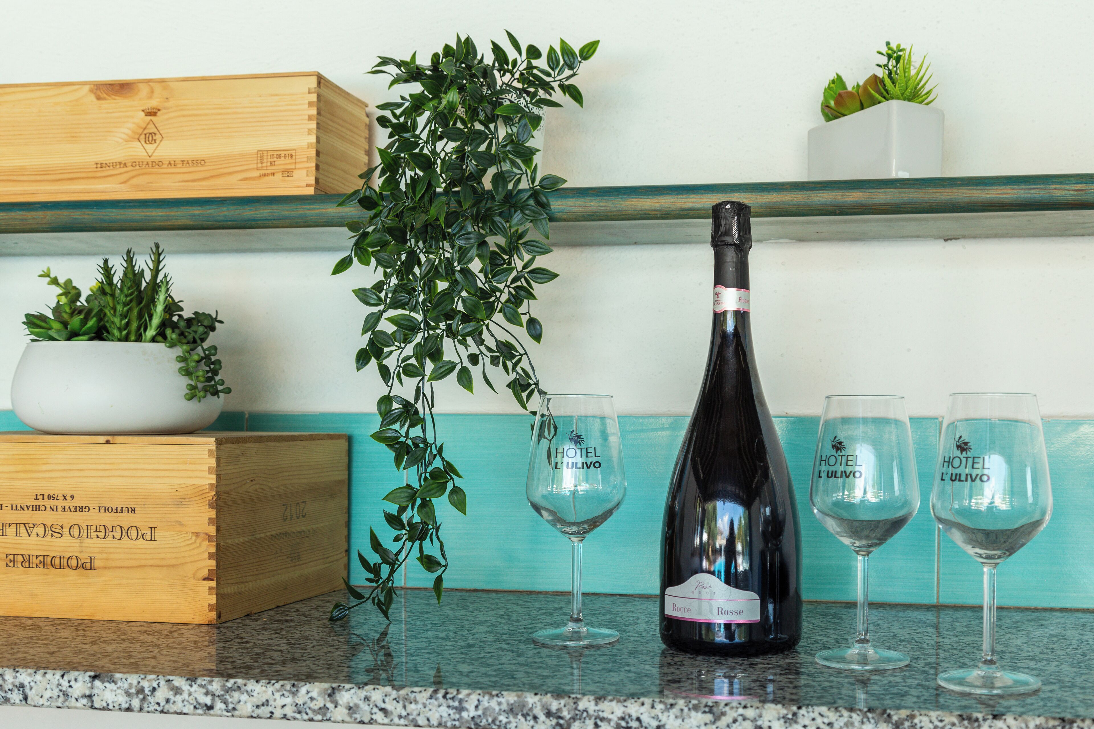 A bottle of wine, two glasses, a wooden crate, and a potted plant on a kitchen counter.