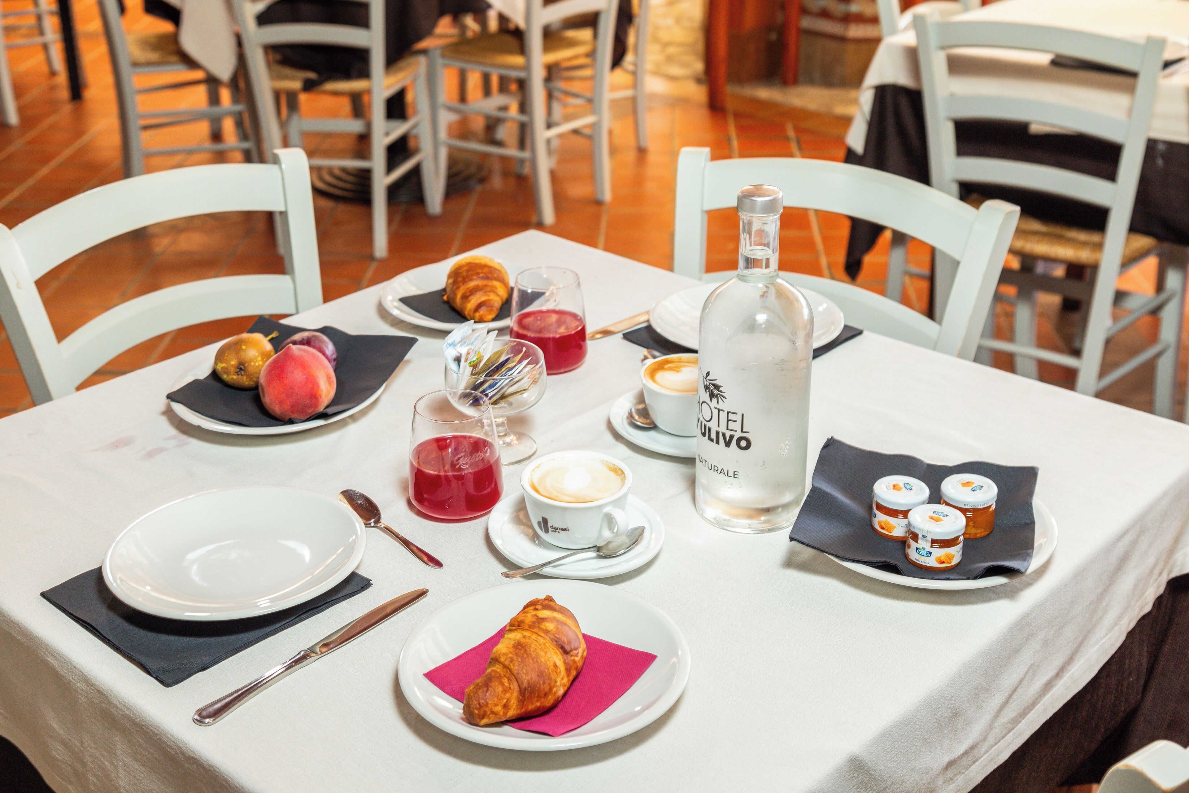 Une table à manger dressée avec des assiettes, des croissants, une bouteille d’huile d’olive et un verre de jus de fruits rouge.