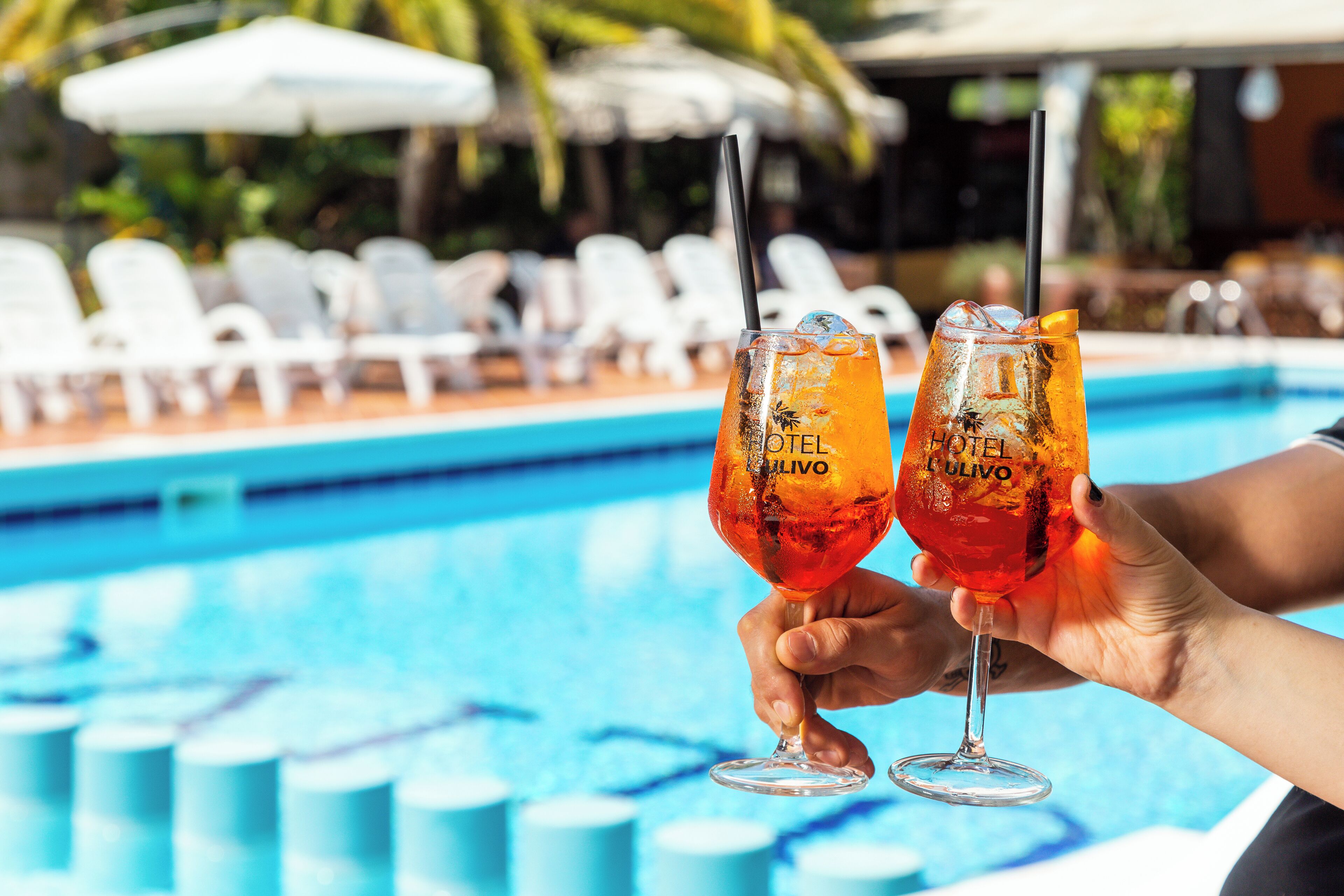 Dos vasos de una bebida roja con pajitas, sostenidos por manos, junto a una piscina con sillas de descanso blancas y sombrillas al fondo.
