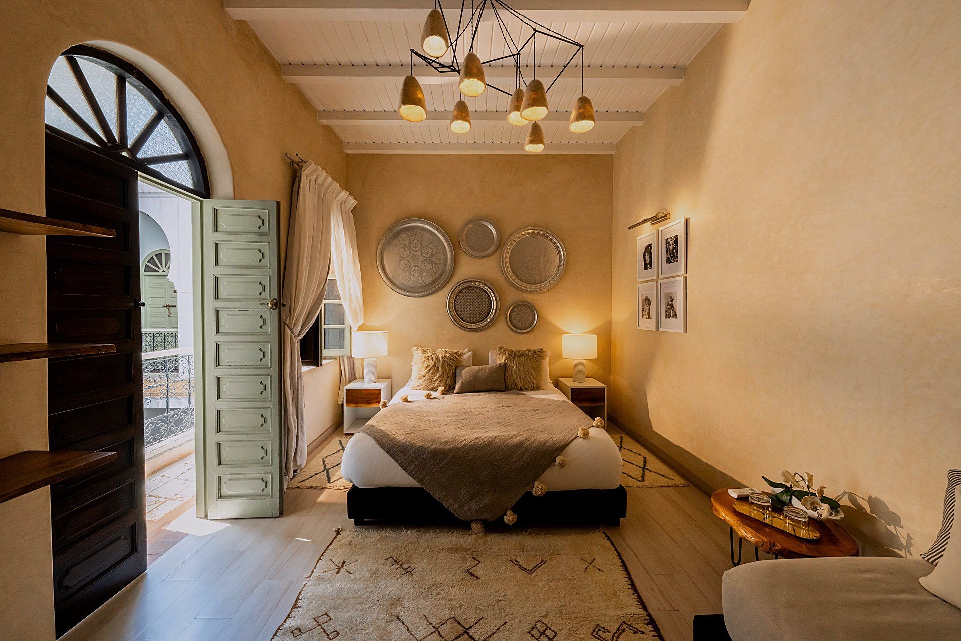 A bedroom with a bed, bedside tables, a chandelier, wall decorations, and a view through an open door.