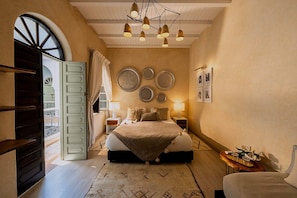 Une chambre à coucher avec un lit, des tables de chevet, un lustre, des décorations murales et une vue sur l’extérieur, visible à travers une porte ouverte.