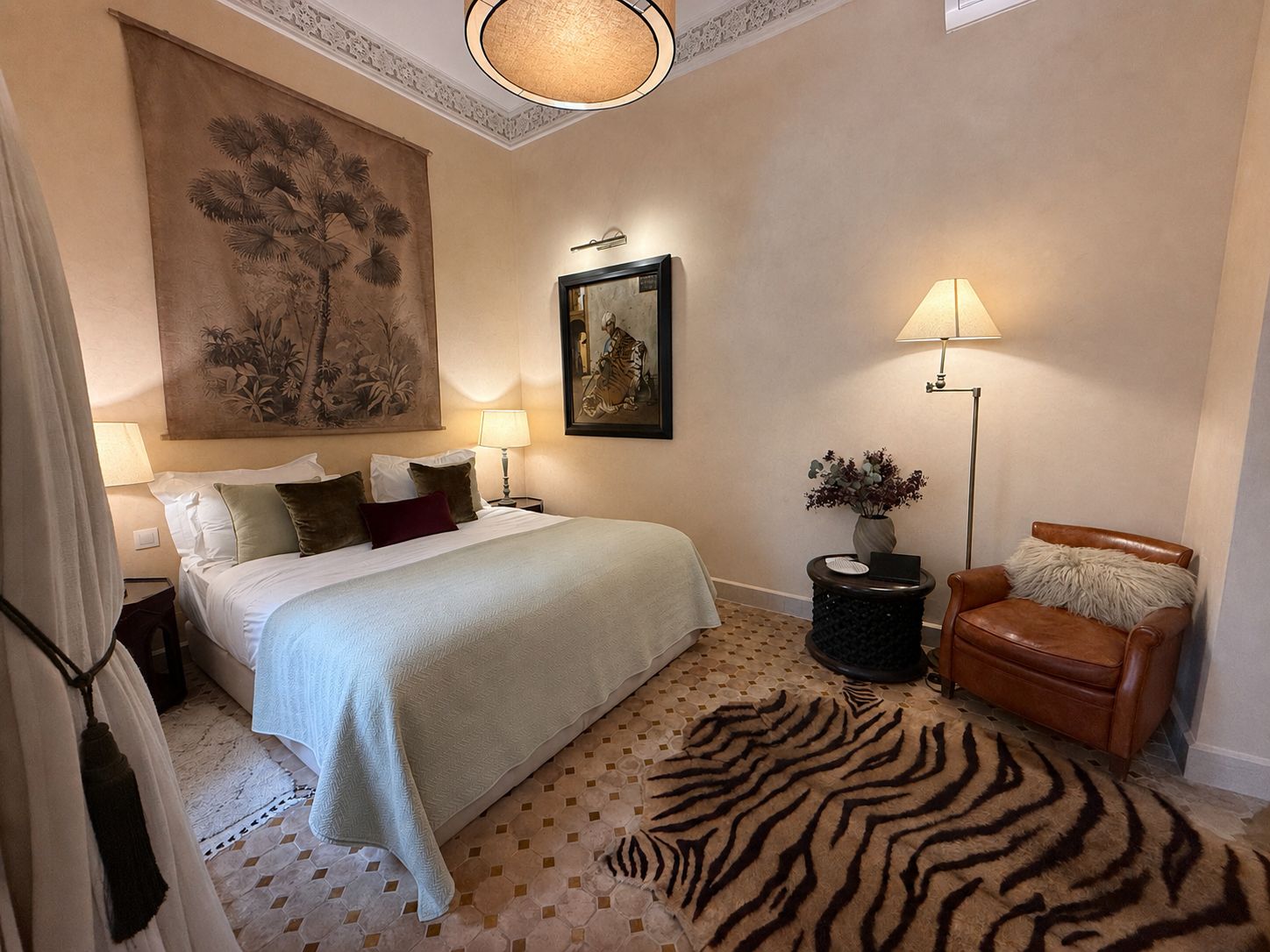 Habitación de hotel con una cama, dos lámparas de noche, un cuadro grande, una obra de arte enmarcada, una silla, una mesita con un florero y una alfombra con estampado de cebra.