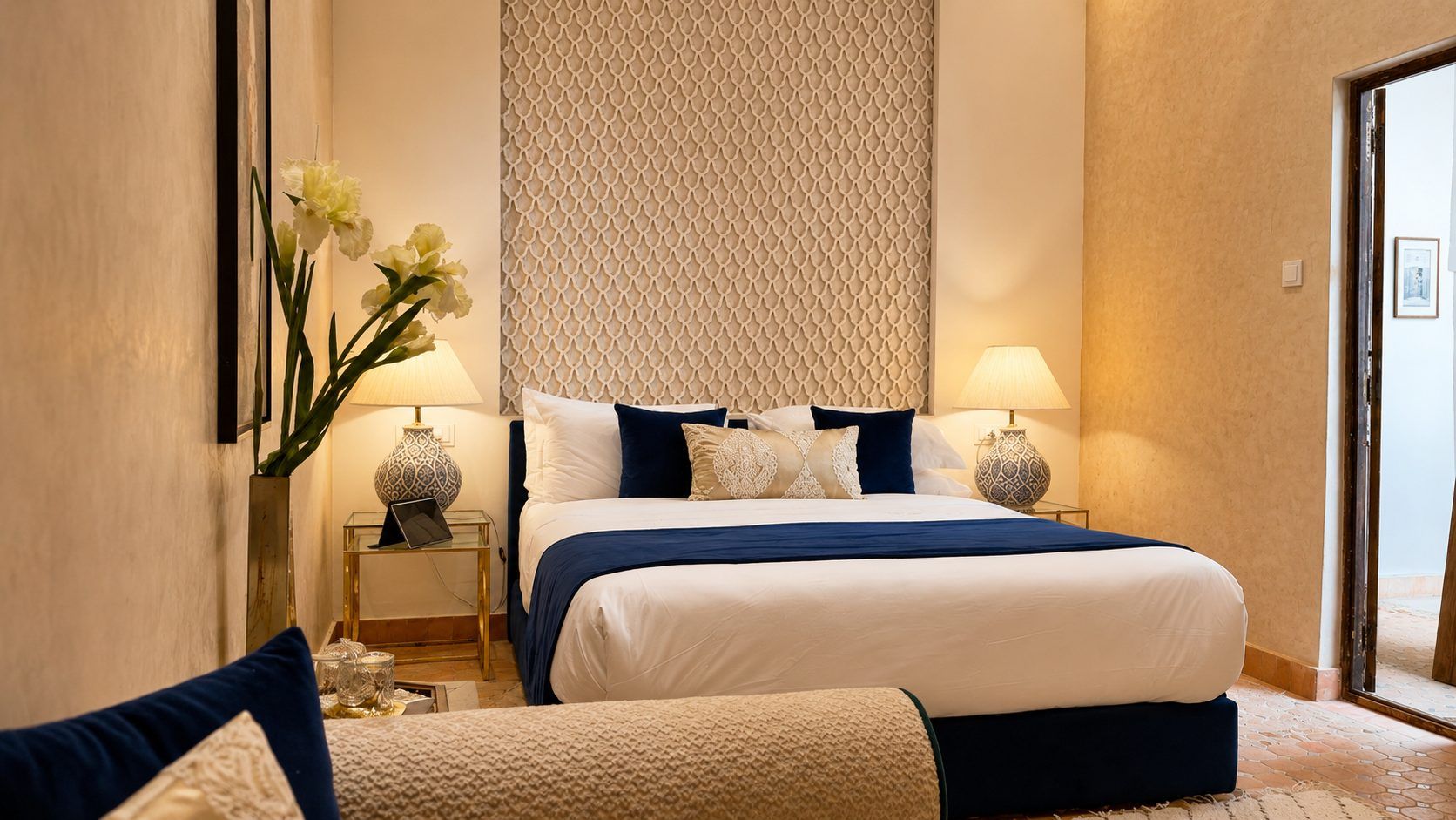 Una habitación de hotel con una cama grande, dos mesitas de noche con lámparas, un jarrón con flores y vistas a través de un ventanal.