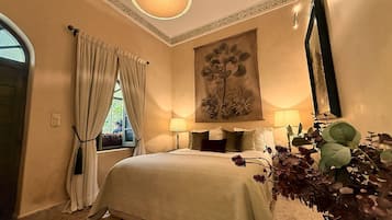 A bedroom with a bed, a window with curtains, a painting on the wall, and a plant.