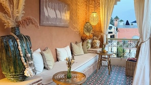 A cozy balcony with a sofa, decorative pillows, a vase with dried flowers, a hanging light fixture, and a view of buildings in the background. - Riad Jona (Marrakech)