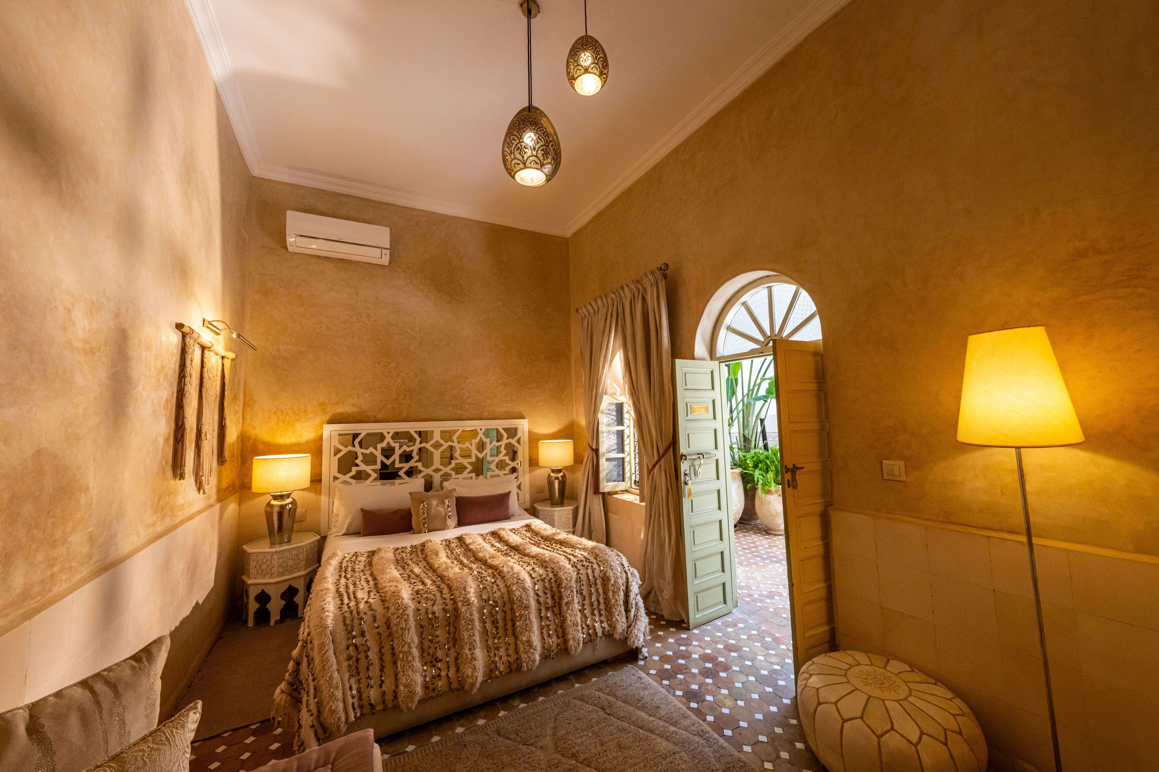 A hotel room with a bed, two bedside lamps, a wall-mounted air conditioner, a patterned rug, and a large window with curtains.
