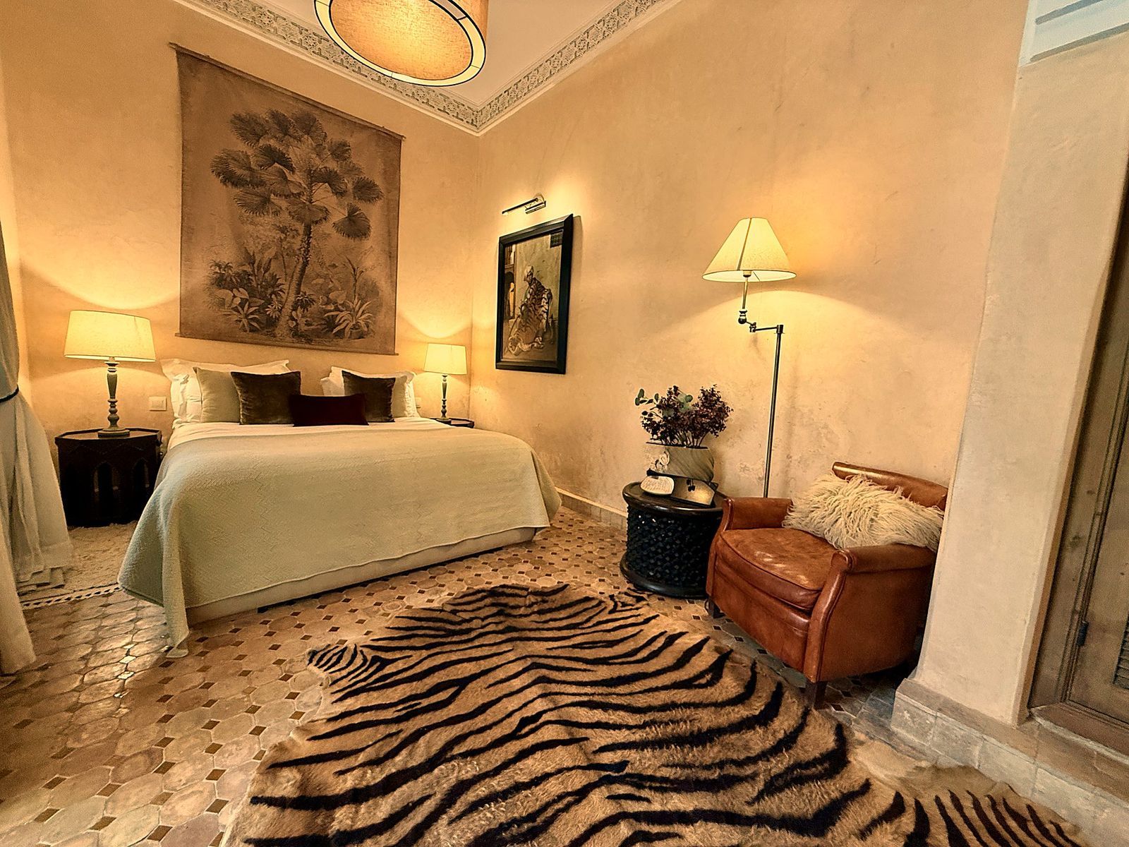 A hotel room with a bed, two bedside tables with lamps, a zebra-patterned rug, a leather armchair, and a large painting on the wall.