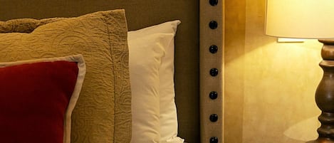 Uma cama bem arrumada com roupa de cama branca e bege, um travesseiro vermelho e uma luminária sobre uma mesinha ao lado.