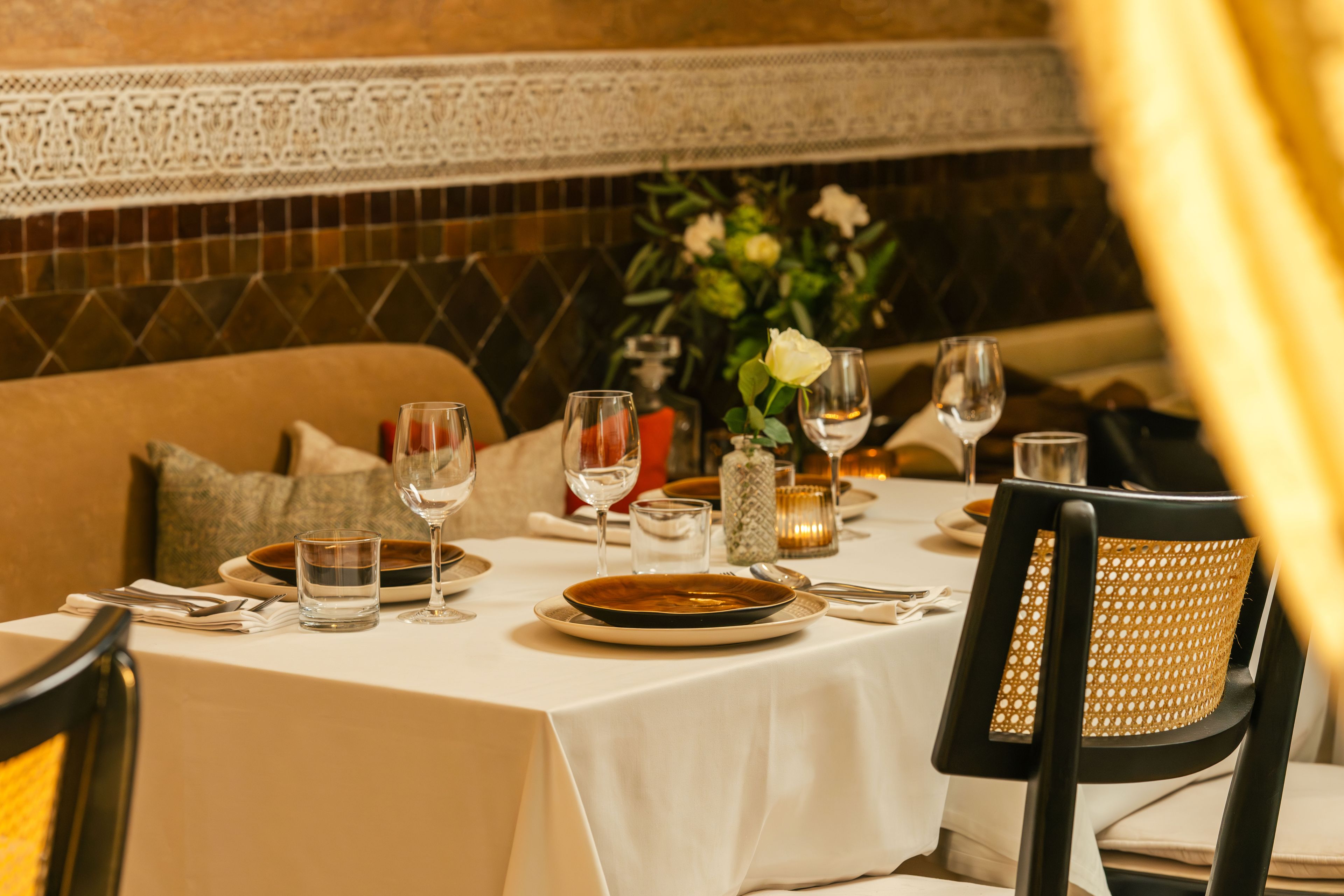 Una mesa lista con platos, vasos y cubiertos, con una silla y un florero con flores al fondo.