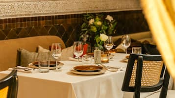 Una mesa lista con platos, vasos y cubiertos, con una silla y un florero con flores al fondo.