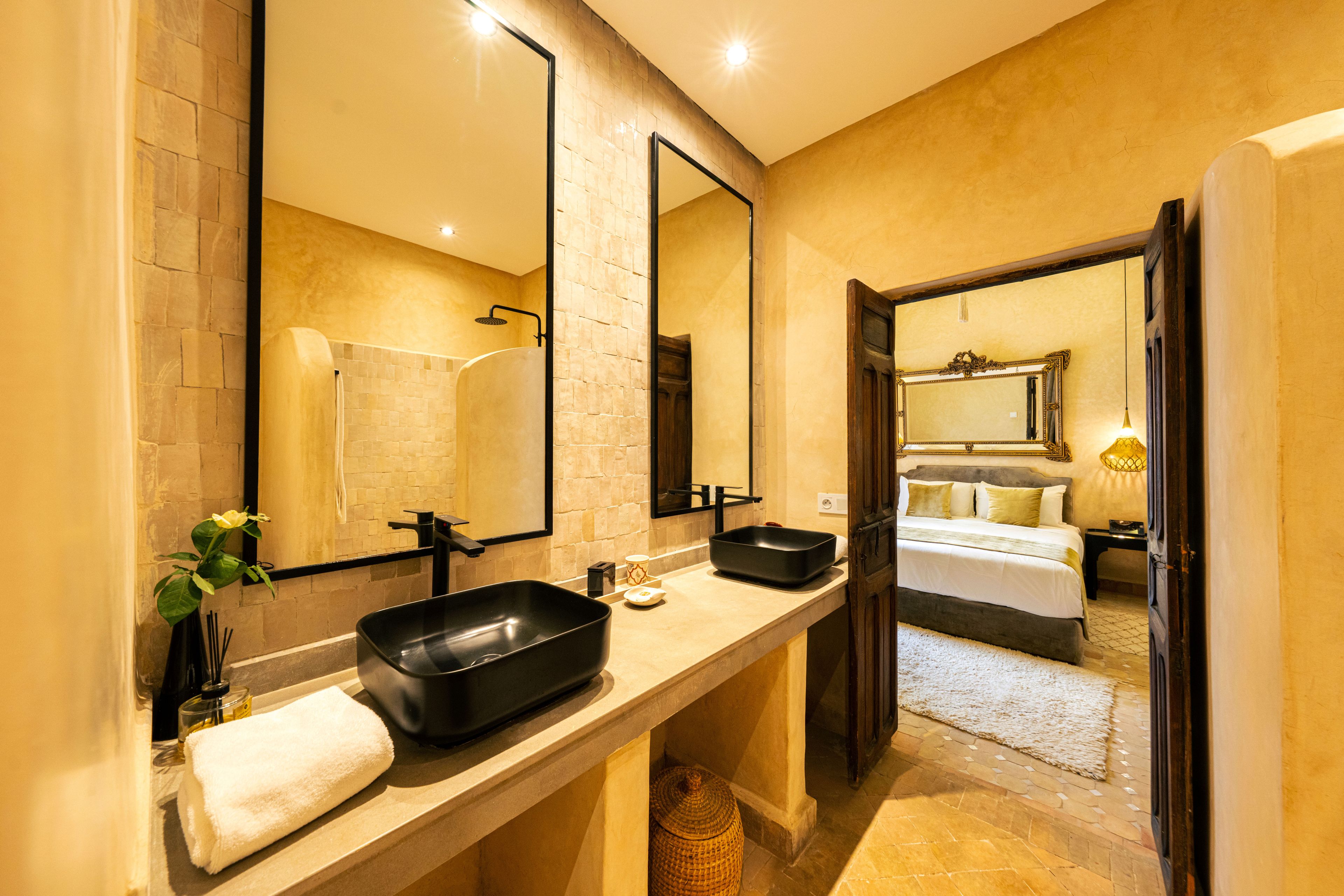 Baño de hotel con un espejo grande, dos lavamanos, una ducha y una habitación visible con cama y mesita de noche.