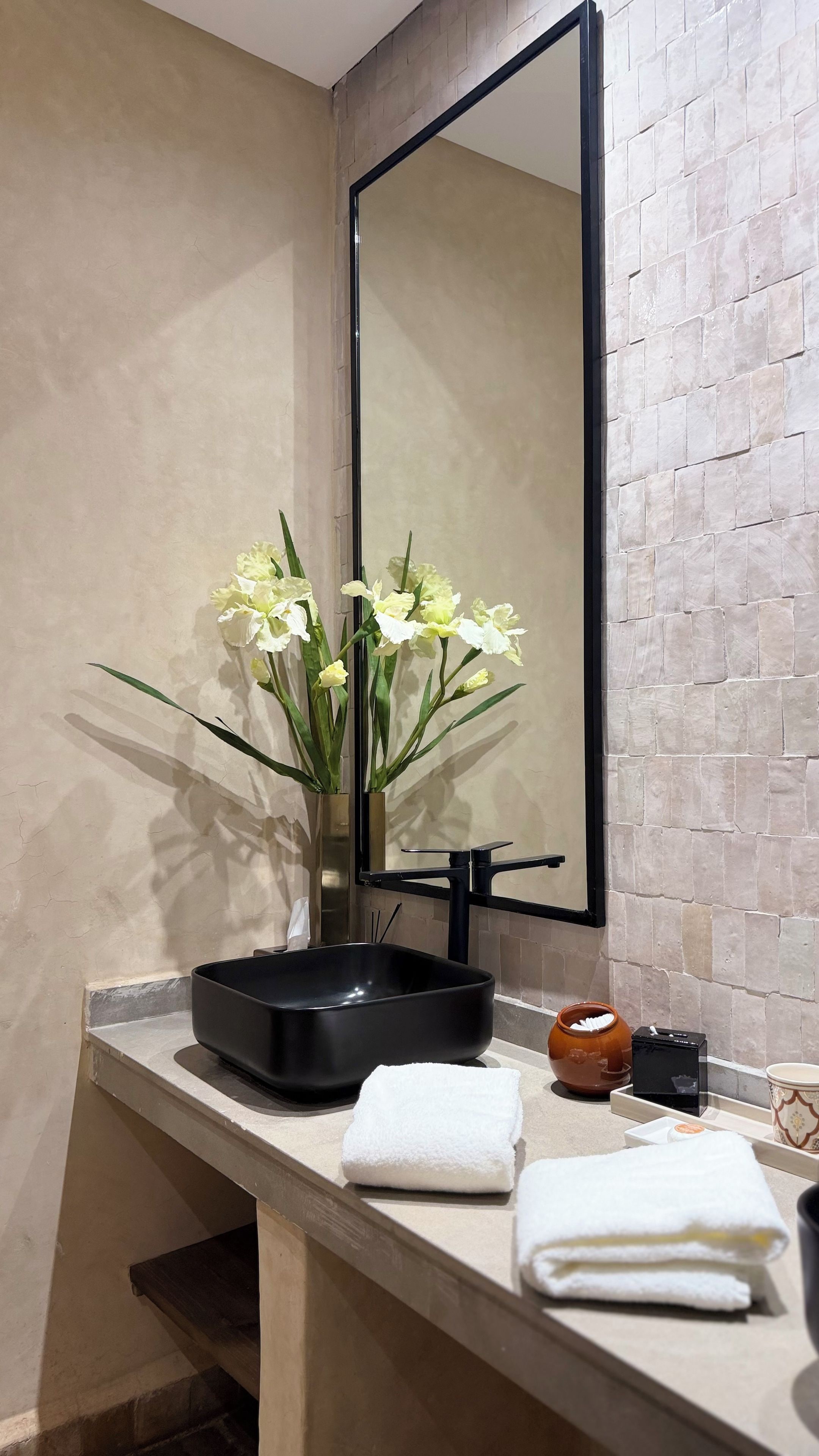 A bathroom with a mirror, a vase with flowers, a black basin, and a towel on the counter.