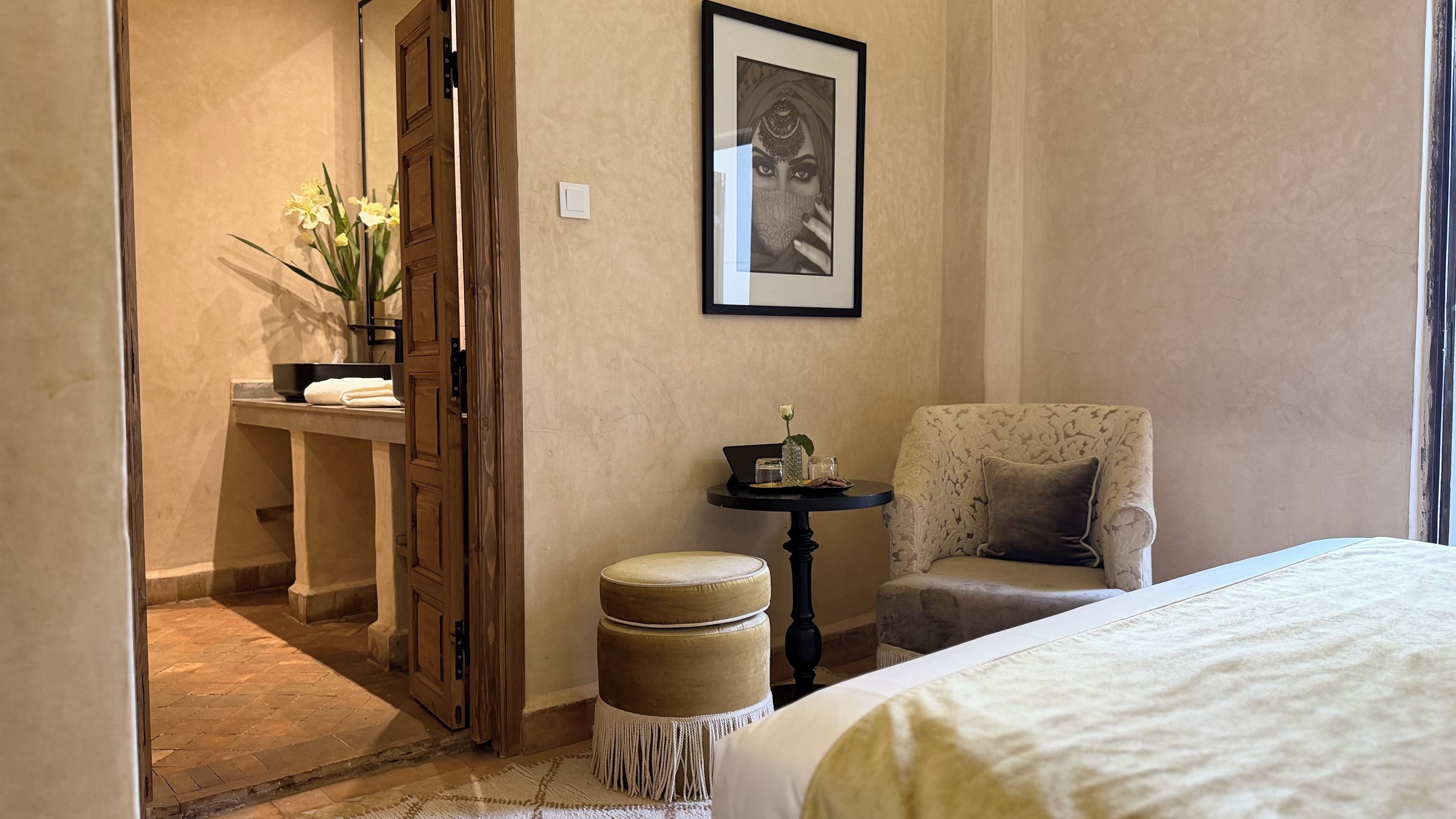 A hotel room with a bed, a chair, a small table with a vase, and a door leading to a bathroom.