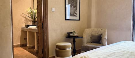 Habitación de hotel con cama, una silla, una mesita con un florero y una puerta que da al baño.