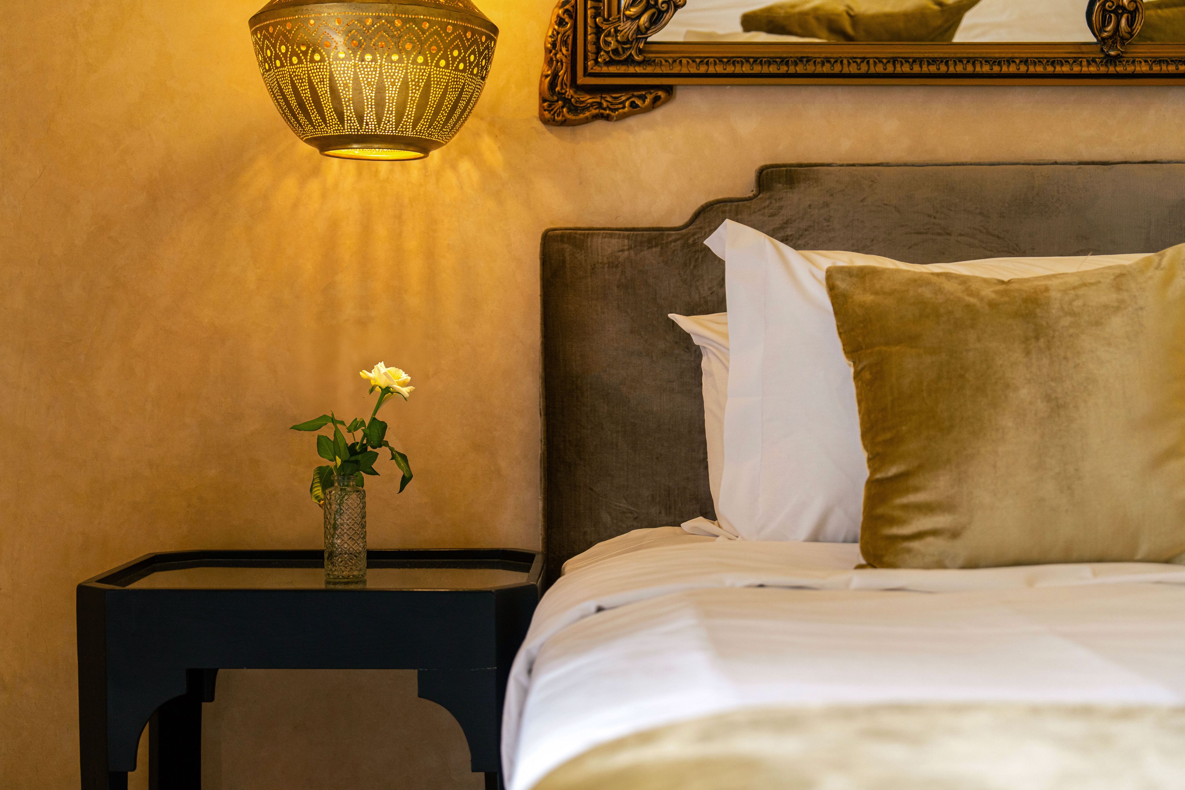 Una cama bien hecha con sábanas blancas y una almohada marrón, un pequeño florero con una sola flor en una mesita de noche, y una lámpara fijada a la pared.