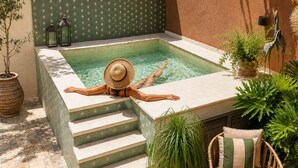 Una persona relajándose en un jacuzzi con escalones que bajan a una piscina, rodeada de plantas en macetas y muebles de mimbre.