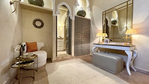 A hotel room with a white sofa, a small round table with a tray, a large mirror, a vanity with a lamp, and a door leading to another room. - Riad Jona (Marrakech)