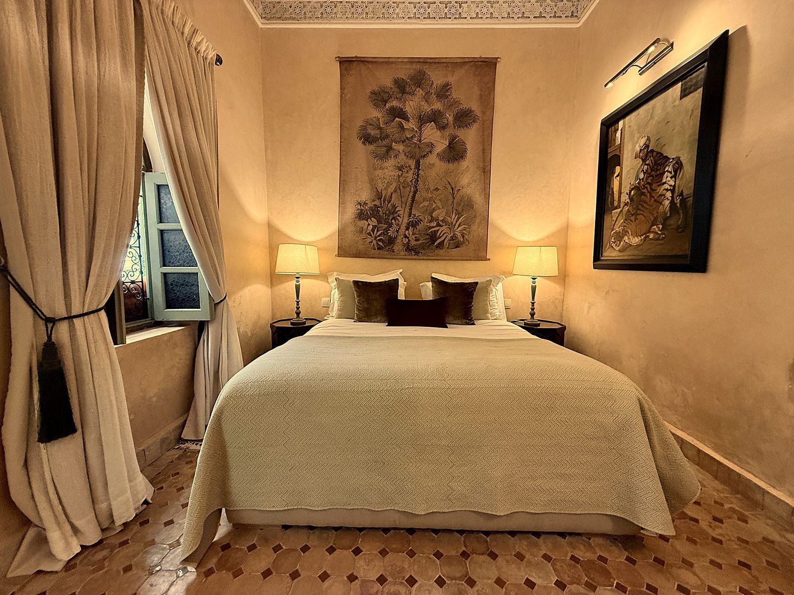 A hotel room with a large bed, two bedside tables, a window with curtains, a wall-mounted painting, and a lamp.
