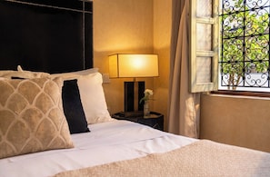Una cama bien hecha con almohadas, una lámpara de noche y una ventana con una reja decorativa de hierro.