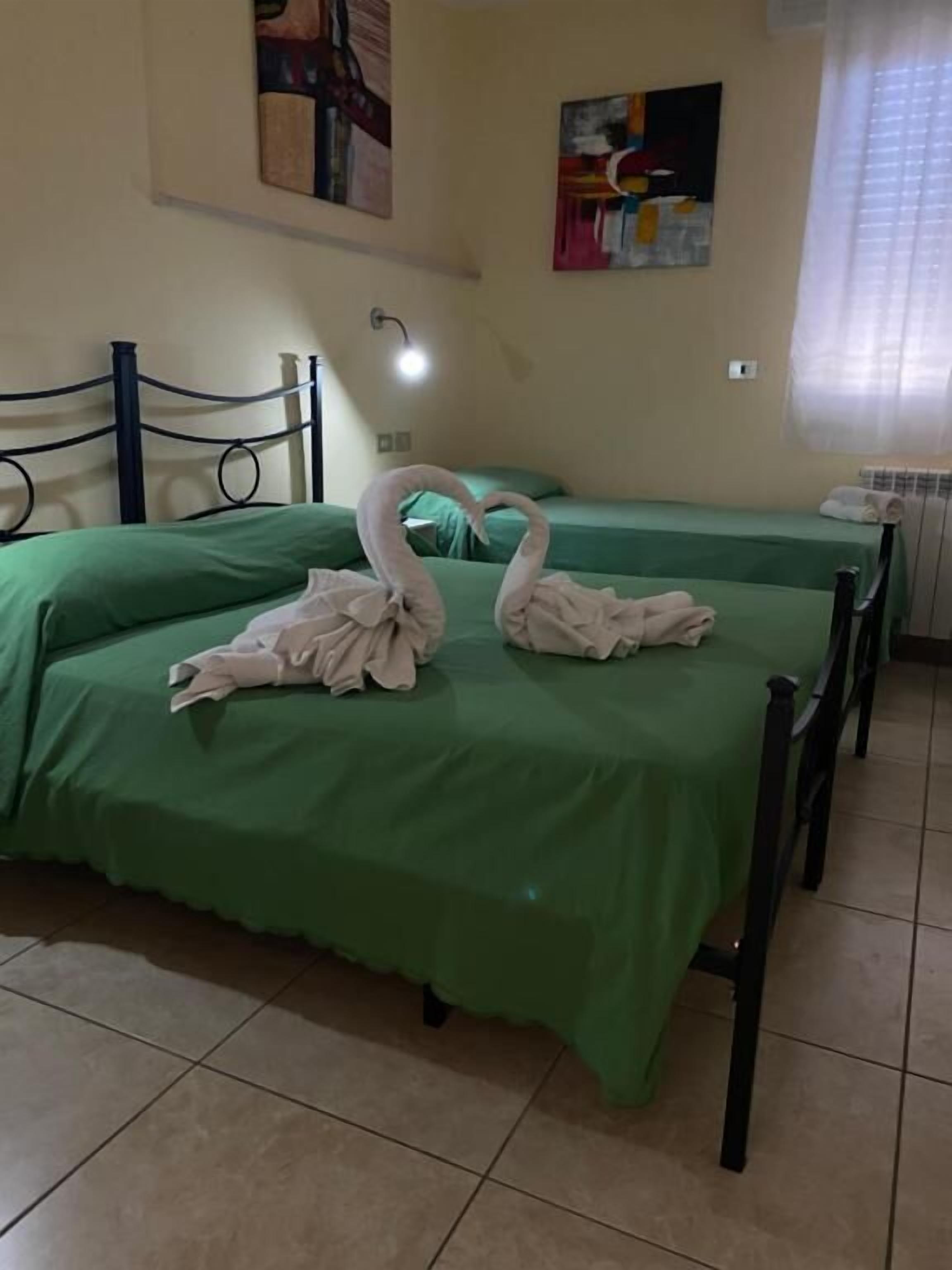 Ein Zimmer mit zwei Betten, jeweils mit grünen Bettdecken bezogen. Über den Betten ist eine wandmontierte Leuchte angebracht.