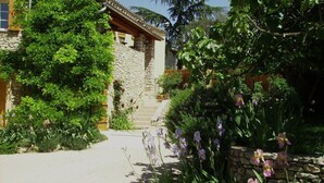 A stone building with a tiled roof, surrounded by lush greenery and flowering plants. - Gîte des Figourières (Sainte-Anastasie)
