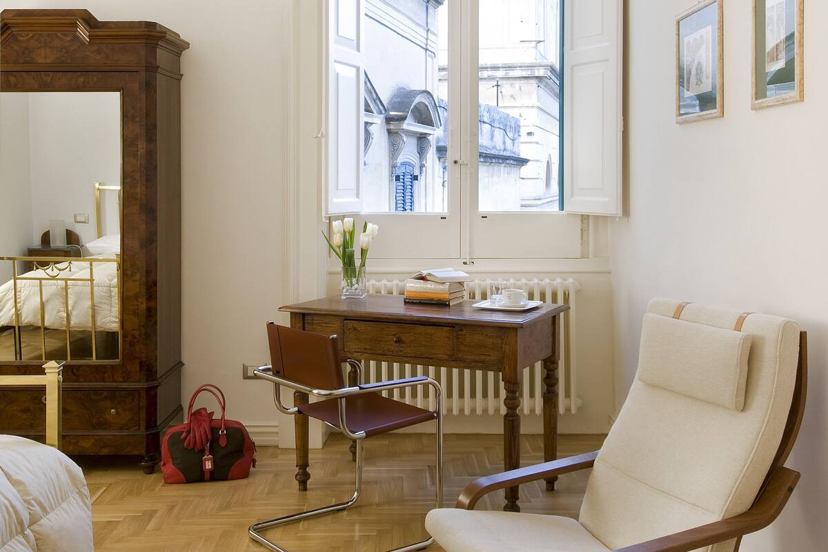 A room with a wooden floor, a large mirror, a wooden desk, a chair, a radiator, and a window with a view of buildings.