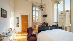 A hotel room with a bed, a desk, a chair, a television, and a view of the city through large windows. - Palazzo De Giorgi (Lecce)