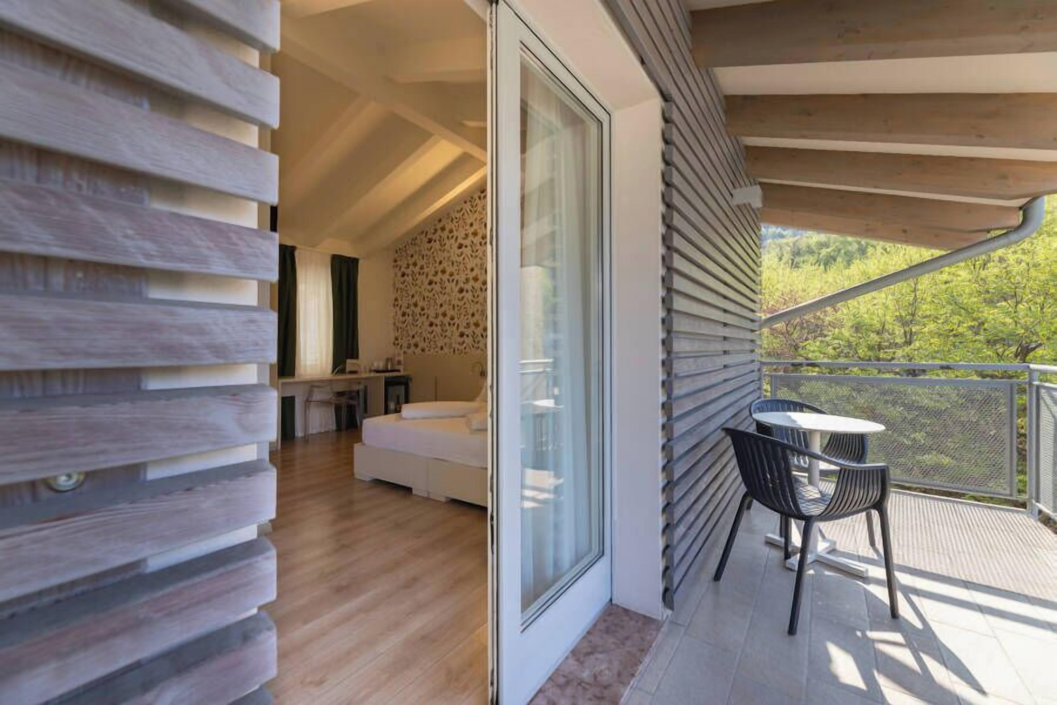 Un balcón con mesa y sillas, una puerta corrediza de vidrio que da a un dormitorio, y vistas al exterior con vegetación.