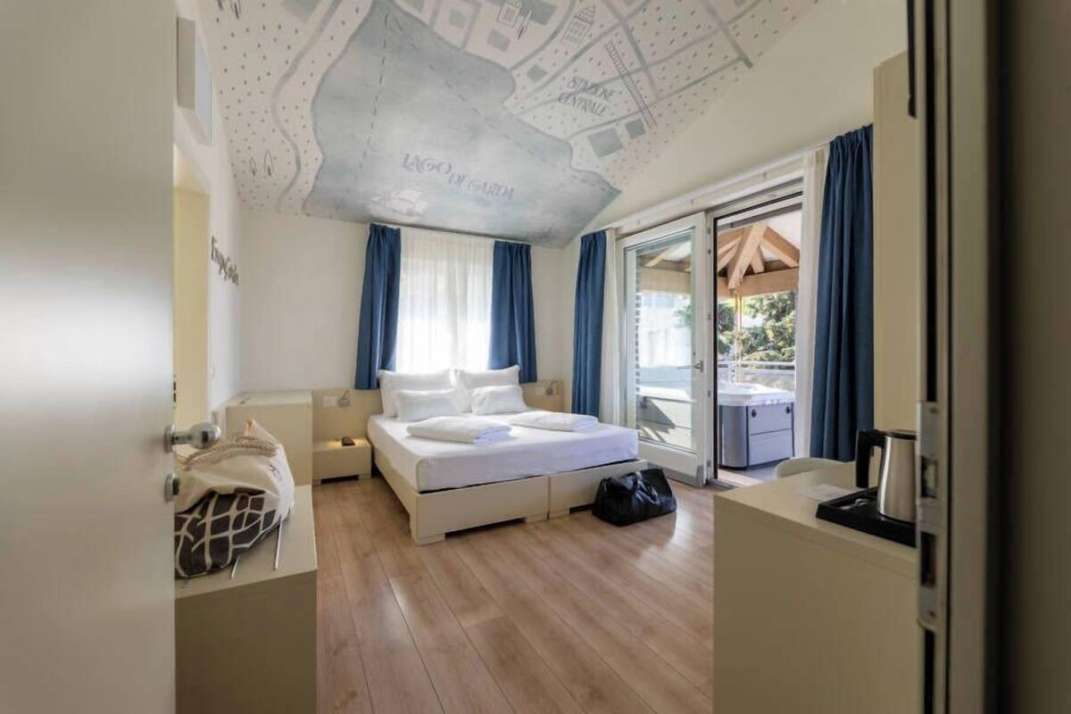 A hotel room with a bed, a desk, a chair, a suitcase, and a view of the outdoors.