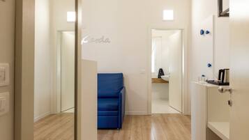 A modern hotel room with a wooden floor, a blue sofa, a glass partition, and a kitchenette area.