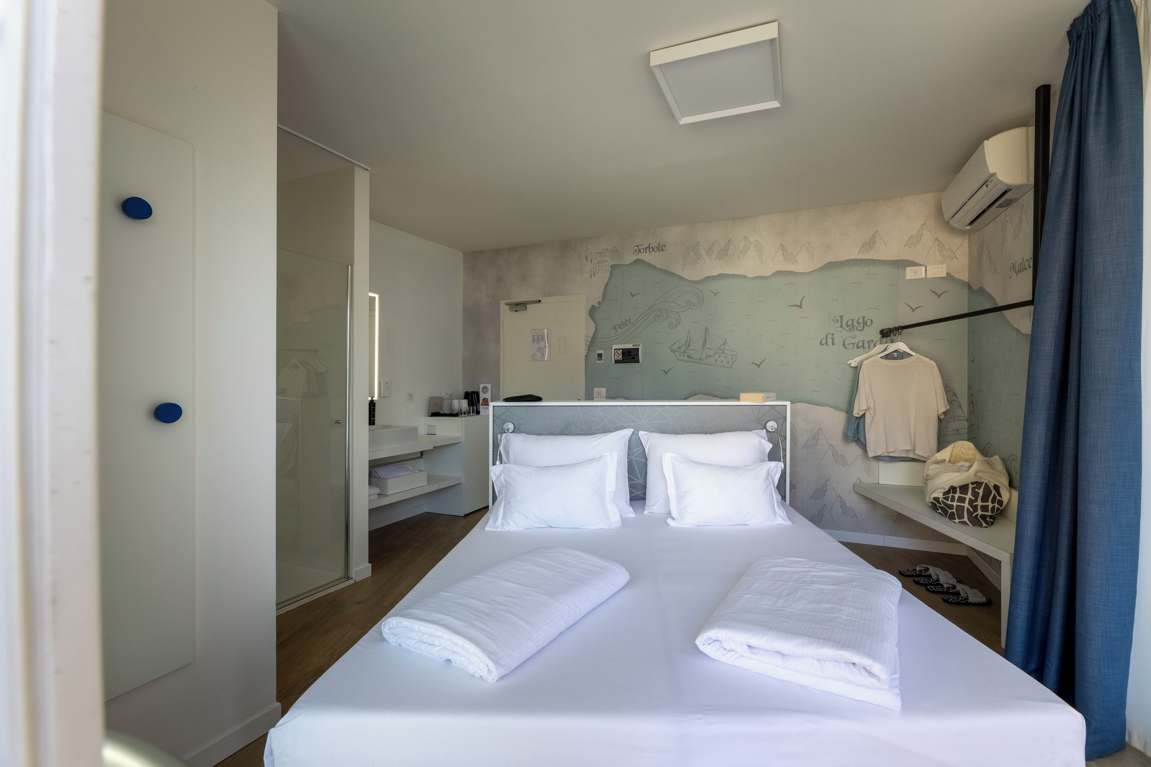 Habitación de hotel con una cama grande, ropa blanca y un baño con cabina de ducha de vidrio, además de un mural en la pared.
