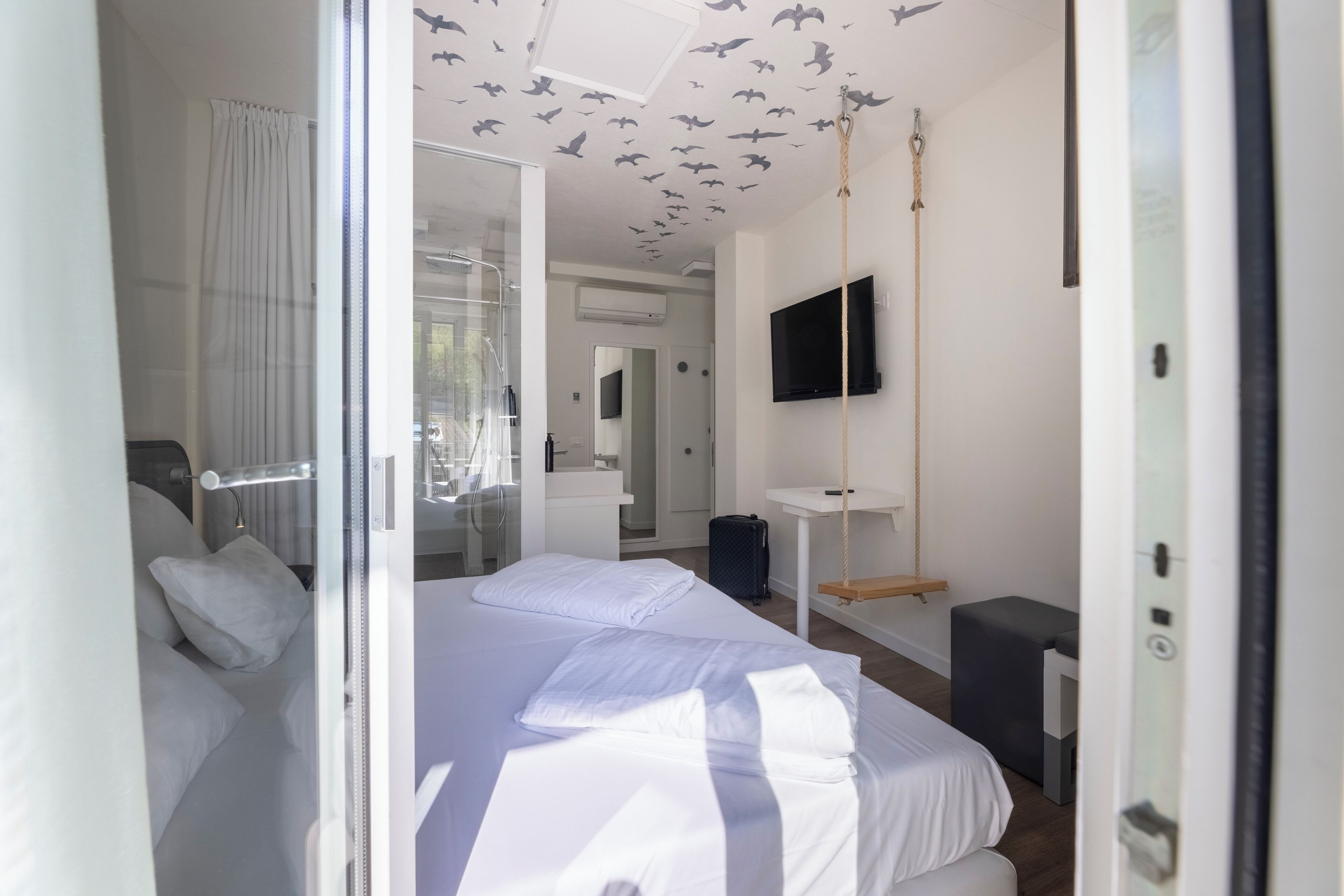 A modern hotel room with a large bed, a TV mounted on the wall, a mirror, and a window with curtains.