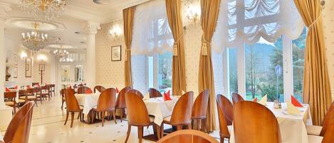A dining area with multiple tables and chairs, large windows with curtains, and chandeliers.