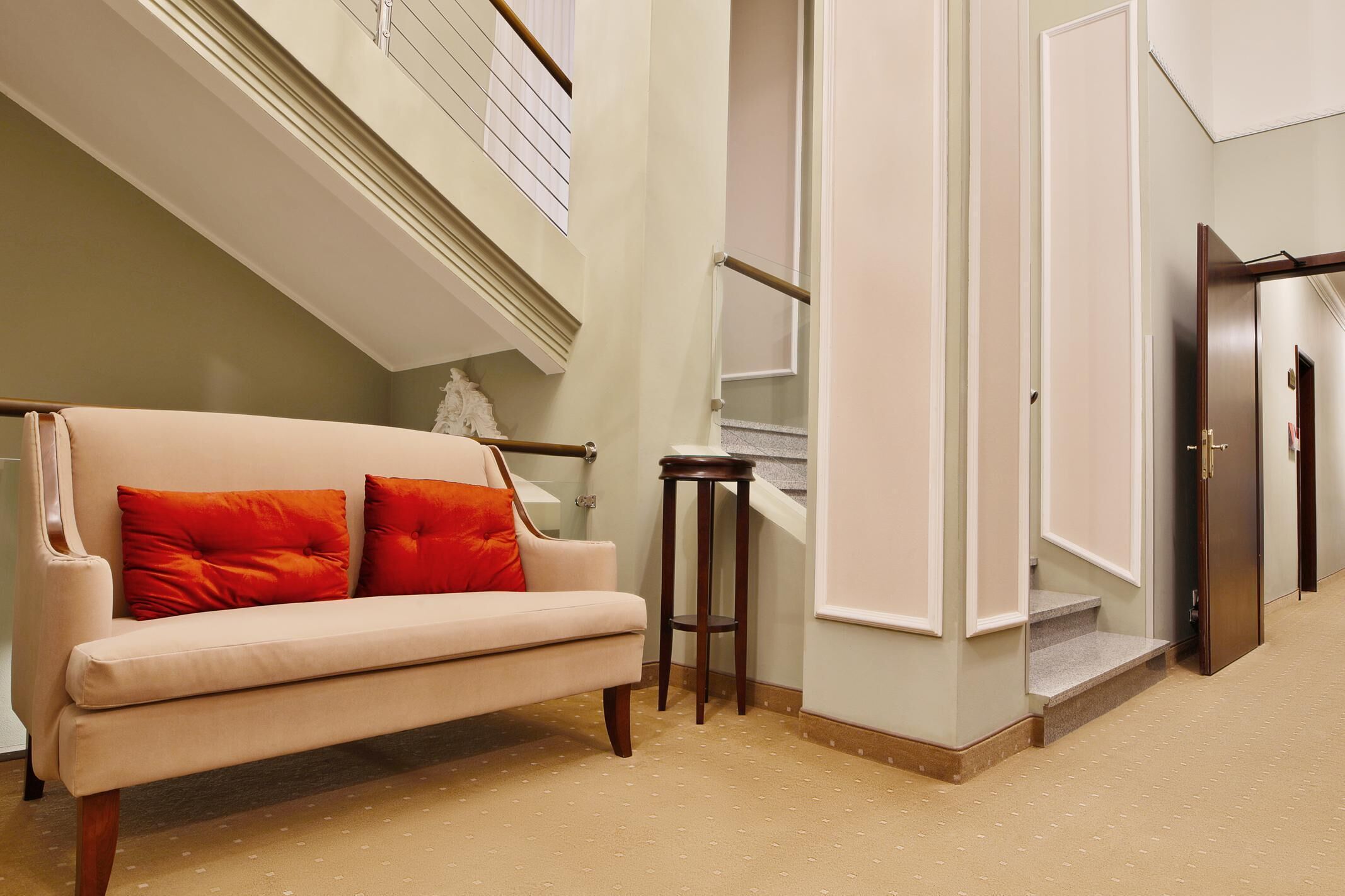 A hallway with a beige sofa, two orange cushions, a wooden stool, and a staircase leading to an upper floor.