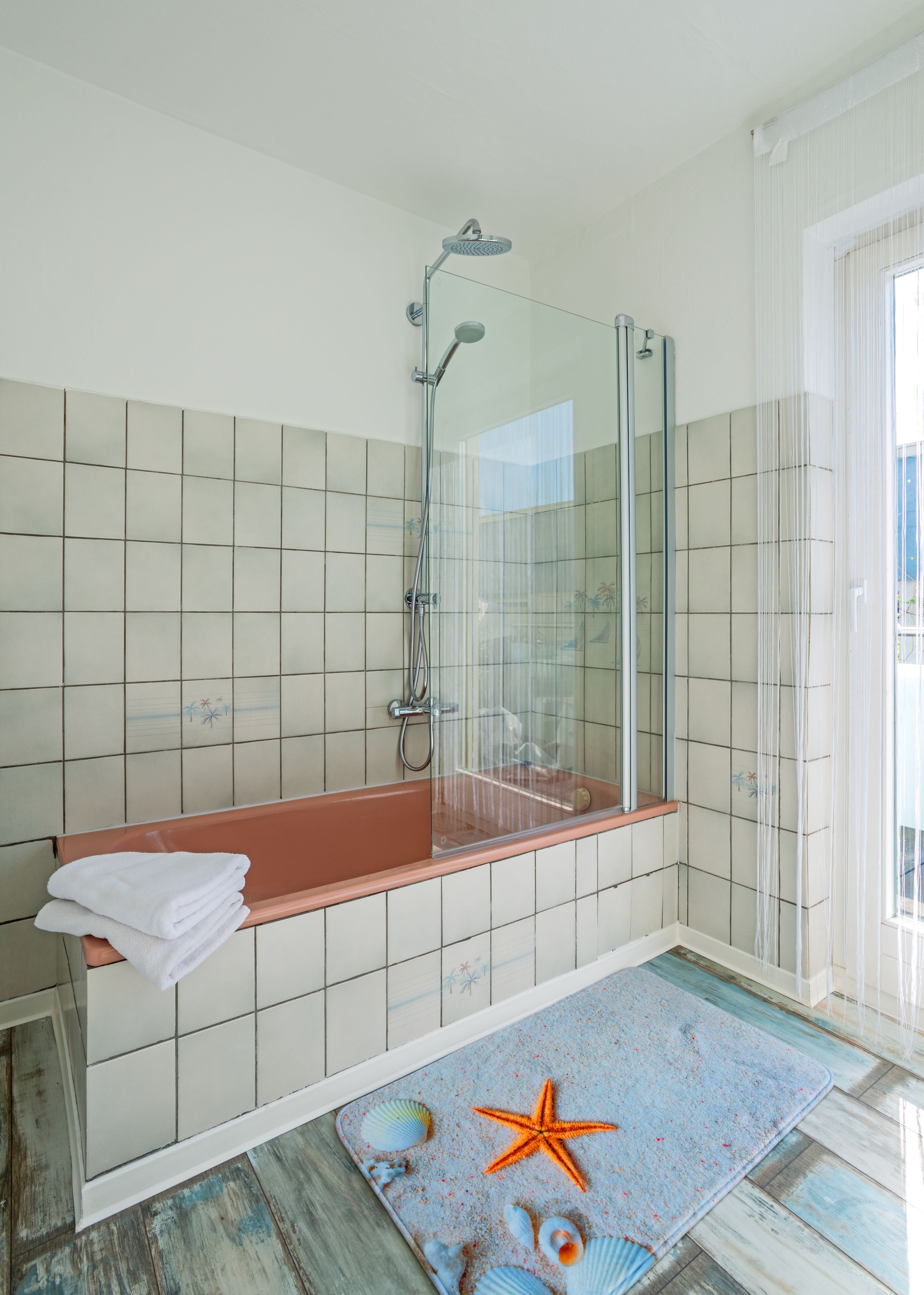 A bathroom with a glass-enclosed shower, a bathtub with a towel, and a decorative rug with sea-themed designs.