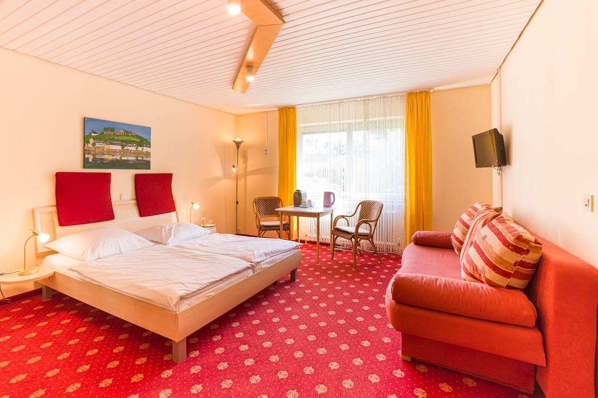 Hotelkamer met een bed, een rode bank, een kleine tafel met stoelen, een schilderij aan de muur en een raam met gordijnen.