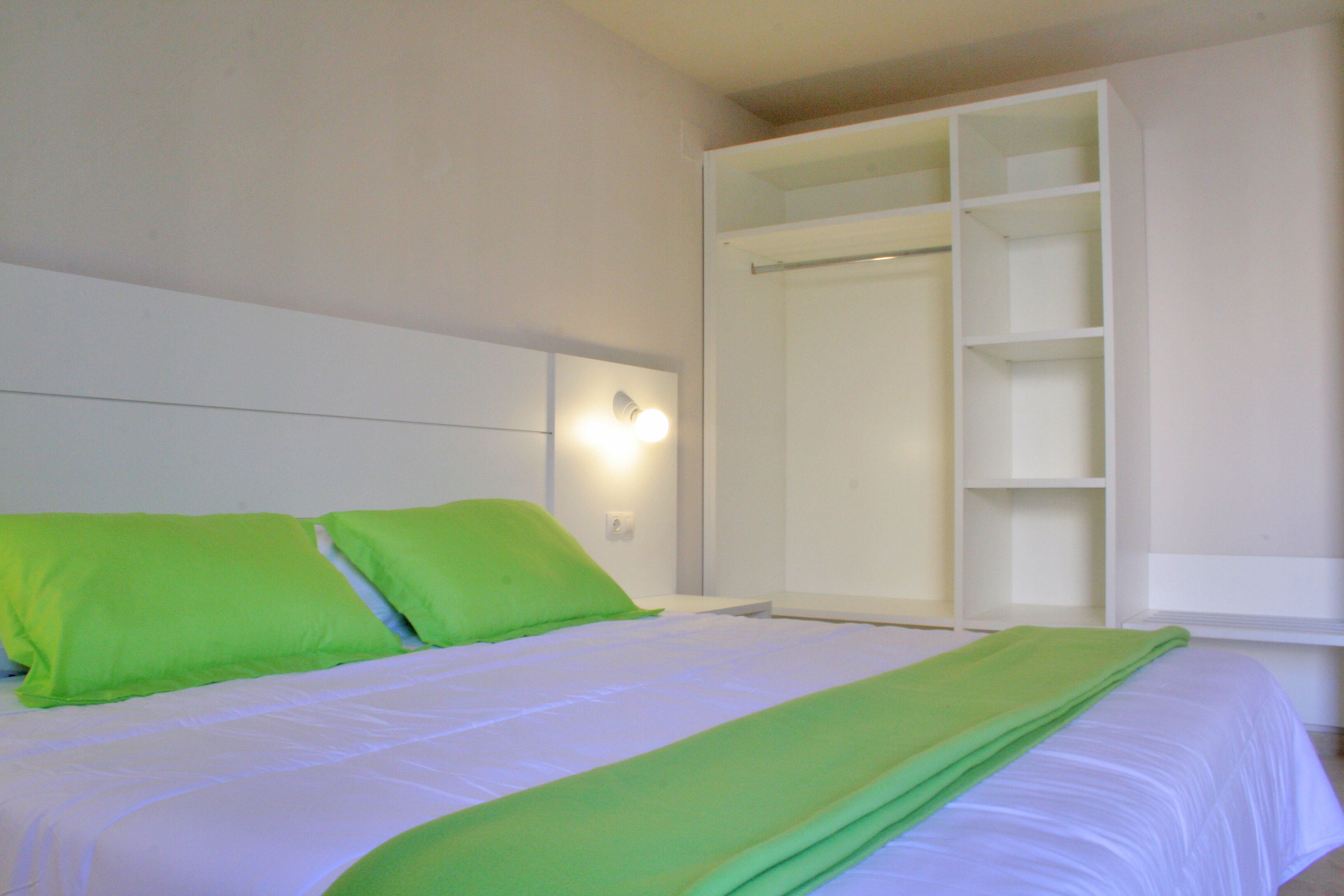 A bed with white bedding and green pillows, a white wardrobe with open doors, and a wall-mounted light fixture.