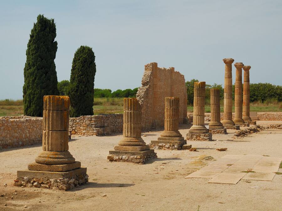 Ancient ruins with stone columns and a stone wall, surrounded by greenery.