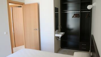 Un dormitorio con una puerta de madera, una cama blanca, un armario negro, un lavabo blanco y una lámpara colgante.