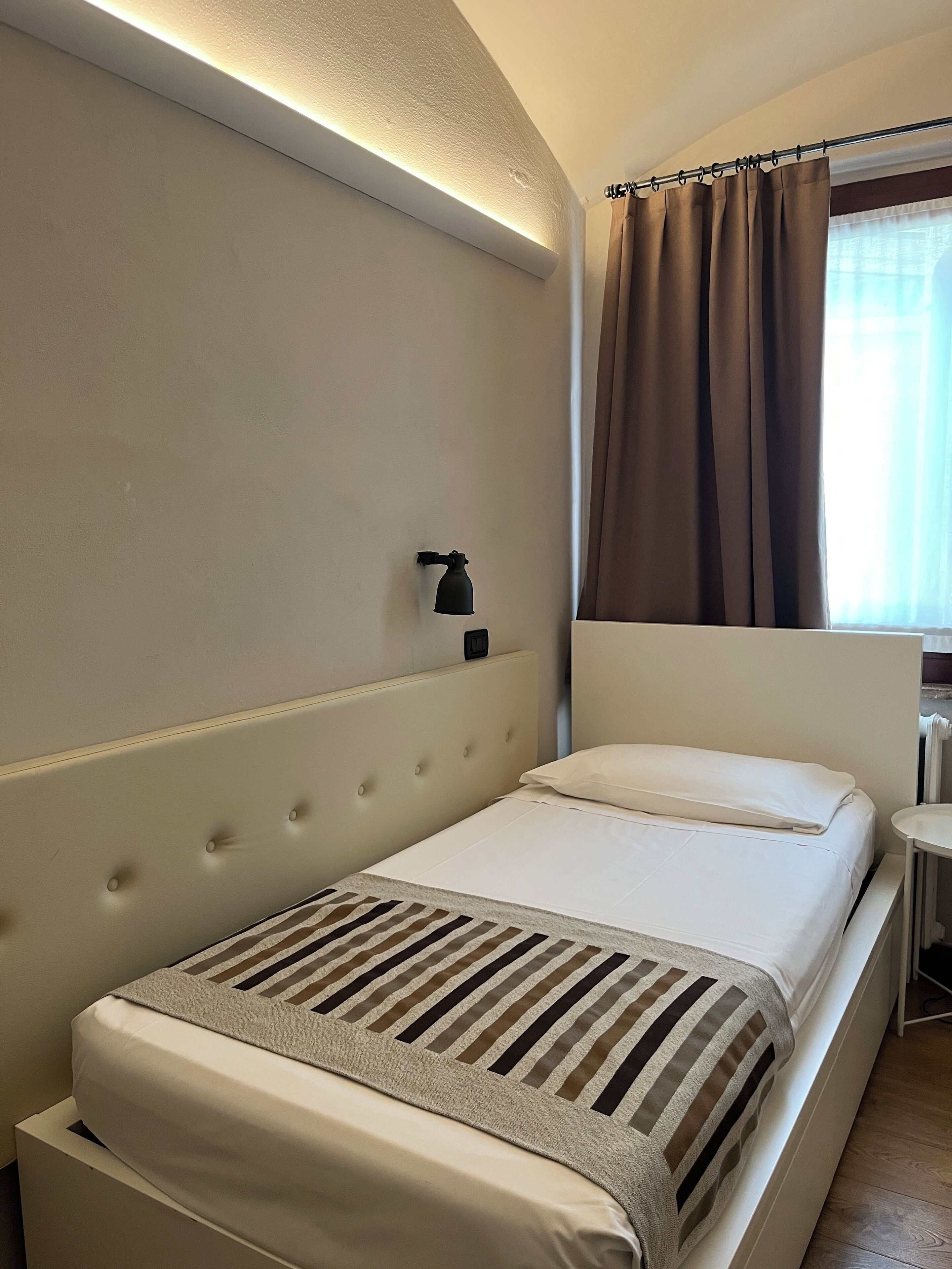 Ein Einzelbett mit einer gestreiften Decke, einer Nachttischlampe und einem Fenster mit Vorhängen.