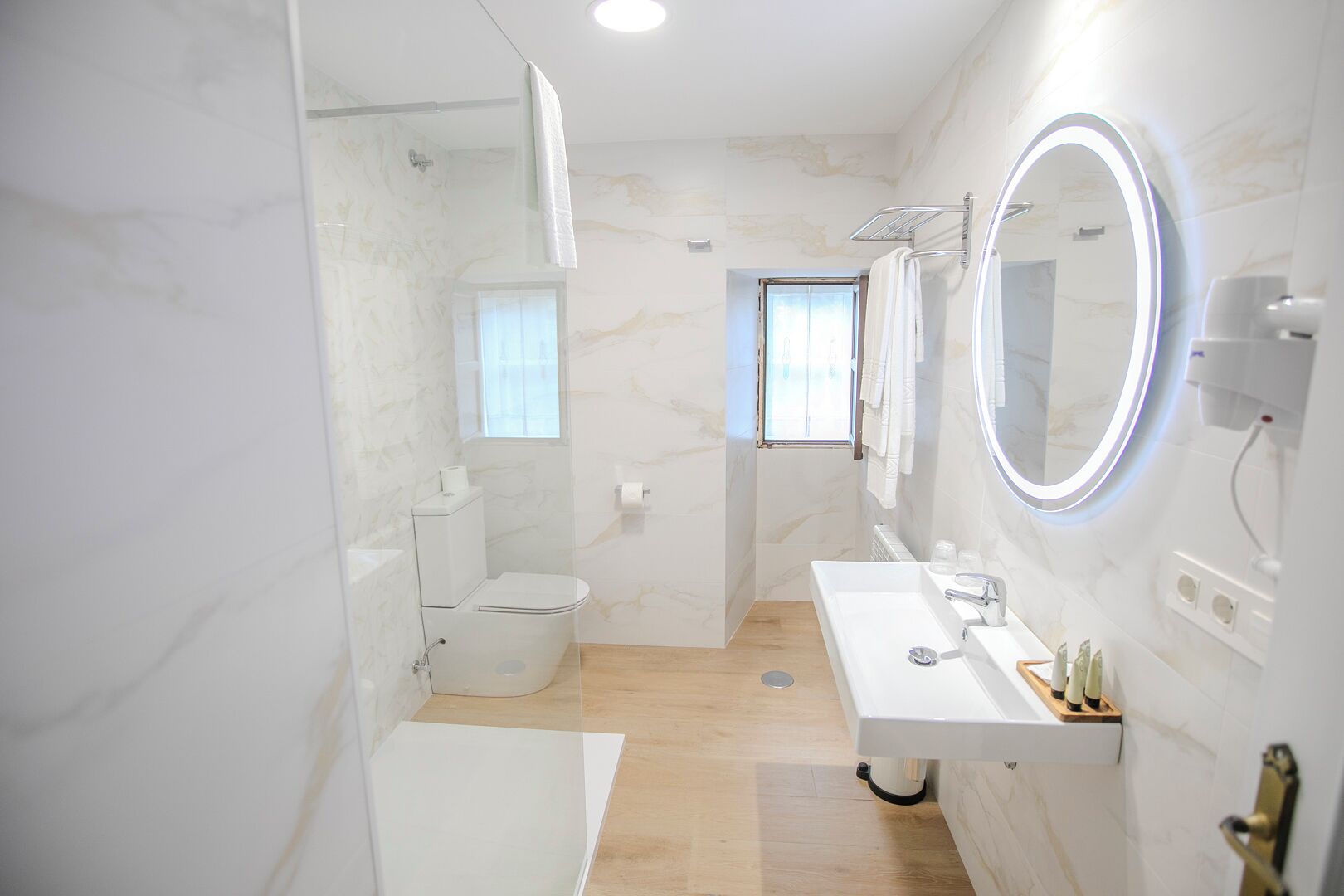 A modern bathroom with a toilet, shower, and sink, featuring marble walls and a wooden floor.
