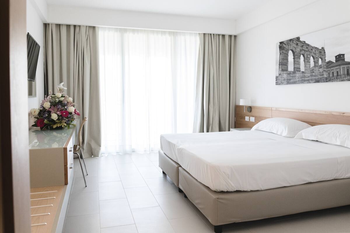 A hotel room with a large bed, a desk with a vase of flowers, a painting on the wall, and a view of the outside through sheer curtains.