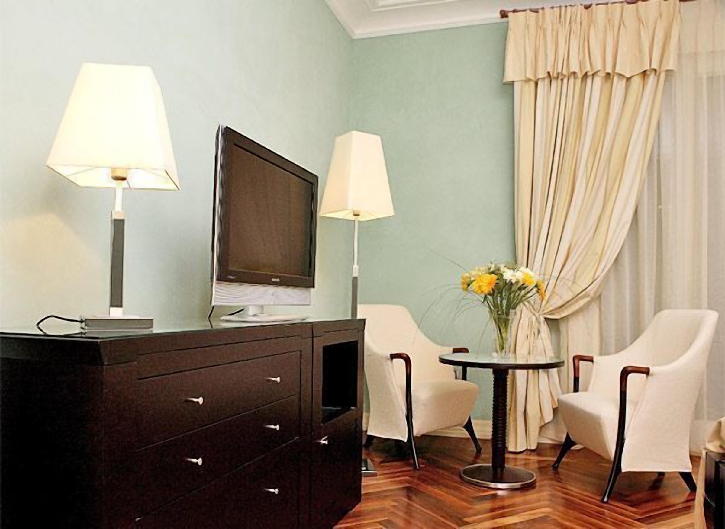 Habitación de hotel con cómoda de madera oscura, dos lámparas, televisor de pantalla plana, pequeña mesa redonda con un florero y dos sillas blancas, junto a un amplio ventanal con cortinas color beige.
