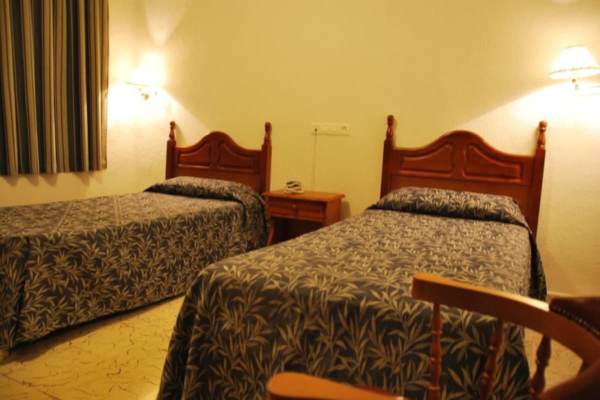 A hotel room with two single beds, wooden headboards, a nightstand, and a chair.