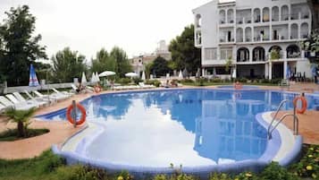 Una piscina con sillas de descanso y sombrillas, rodeada de vegetación y un edificio con varios balcones.