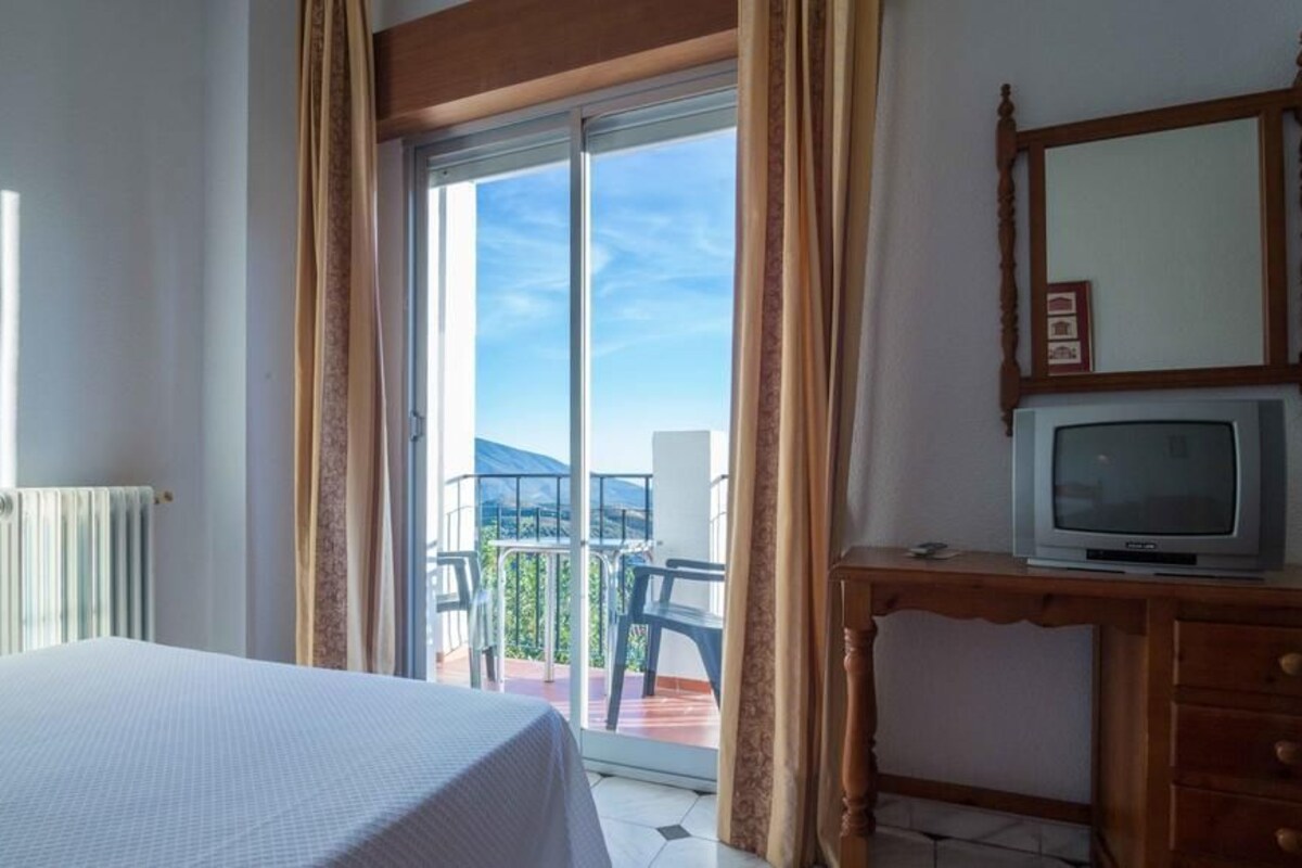 A hotel room with a bed, a TV on a wooden dresser, a balcony with chairs, and a view of the mountains.