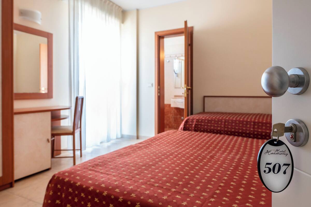A hotel room with a bed, a desk, a chair, a mirror, and a door with a keycard lock.